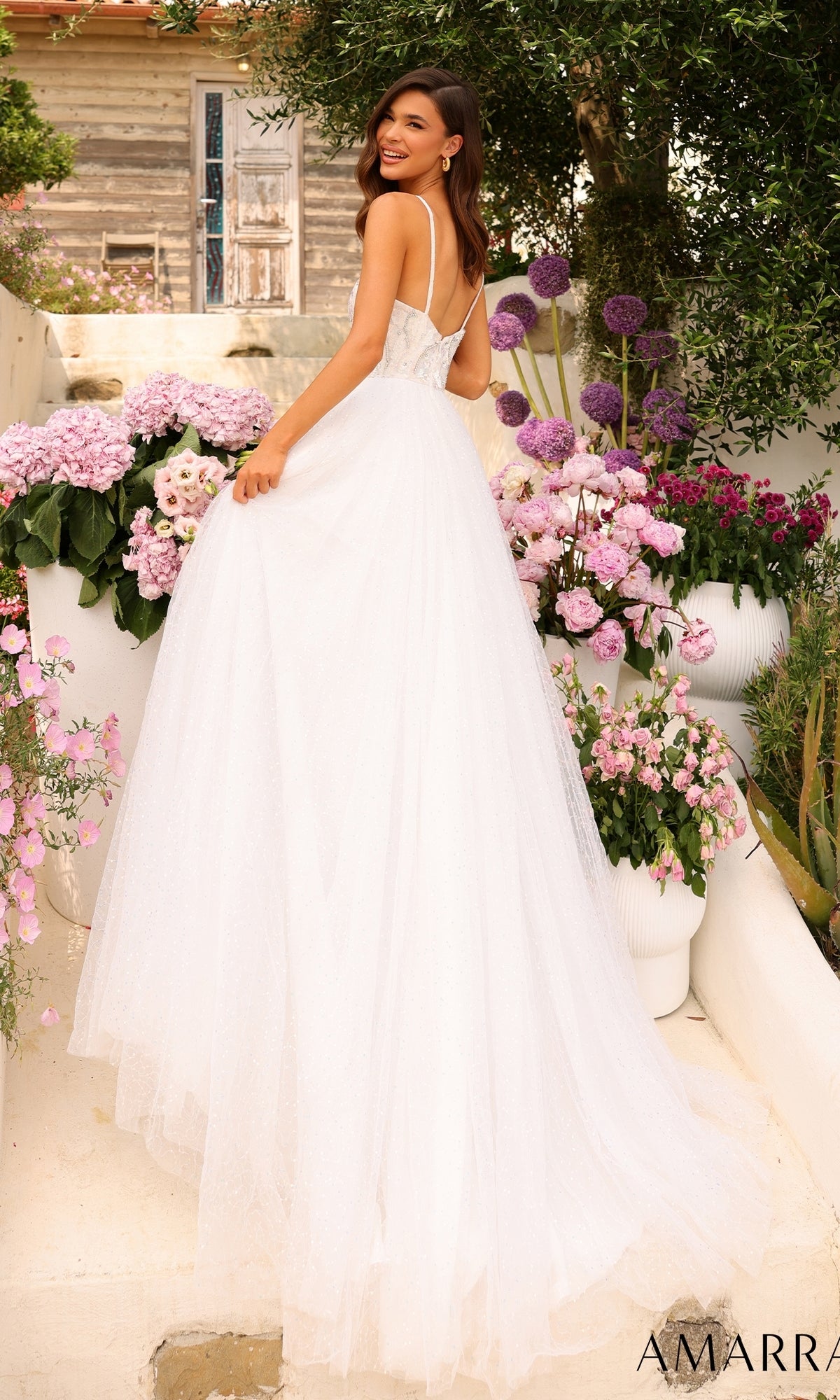 A woman wearing the Amarra Sequin-Bodice Long A-Line Prom Dress 94020 stands outside among pink and purple flowers, smiling over her shoulder.